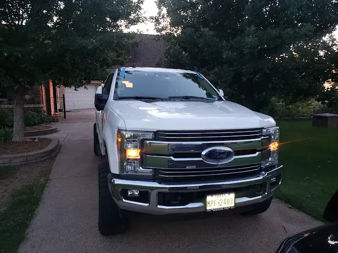 White Ford pickup truck parked on a driveway with a house and trees in the background. The truck's blinker is on.