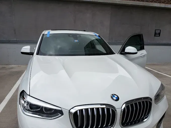 White BMW SUV parked with driver's side door open, in a parking lot, gray wall in background.