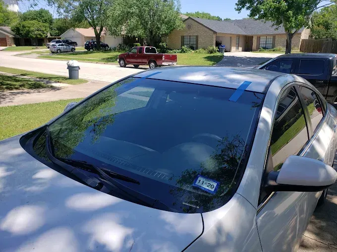 White car with blue tape on windshield, parked in front of a house on a sunny day.
