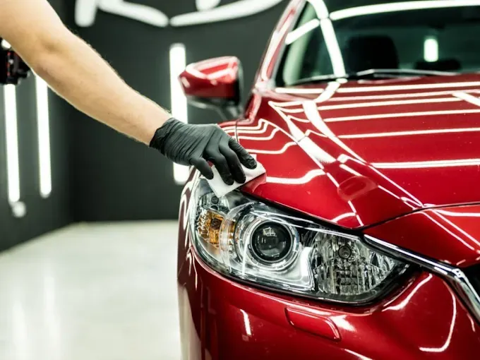Person applying wax to a red car, wearing black gloves. Car is in a brightly lit garage.