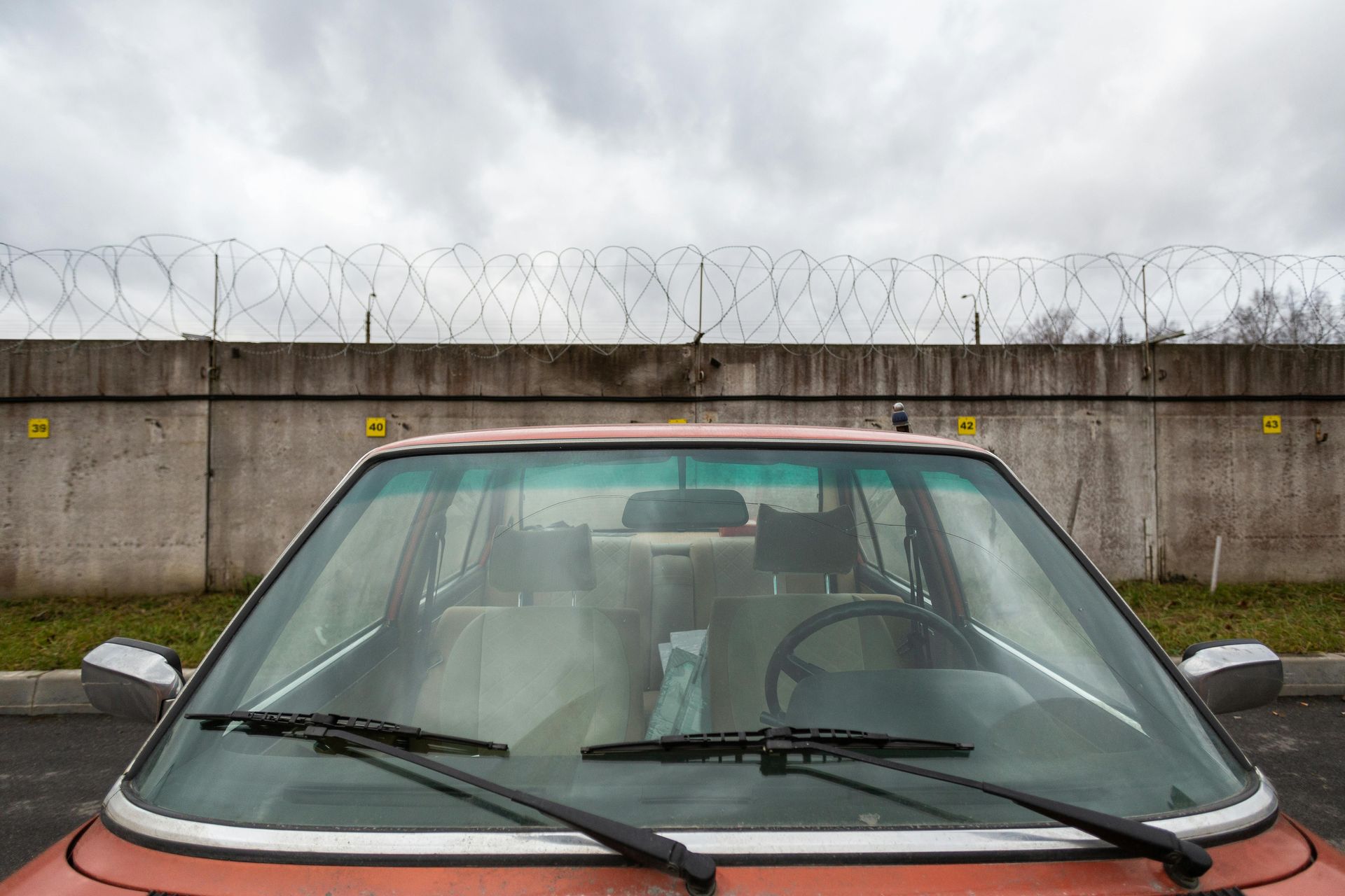Red car parked in front of a concrete wall topped with barbed wire, under a cloudy sky.