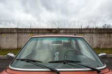 Red car parked in front of a concrete wall topped with barbed wire, under a cloudy sky.