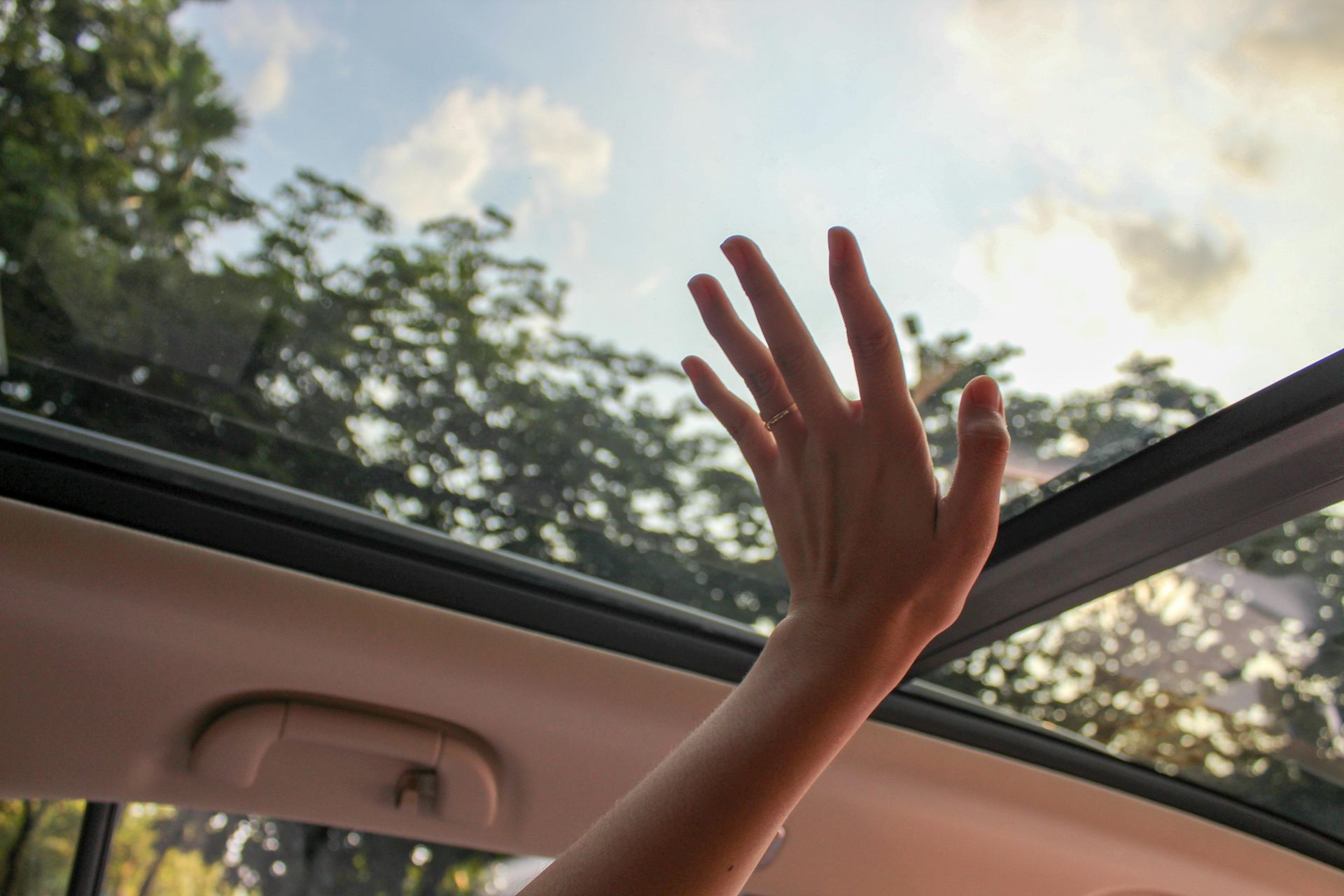 Hand reaching up toward the sky through an open car sunroof, with trees visible in the background.