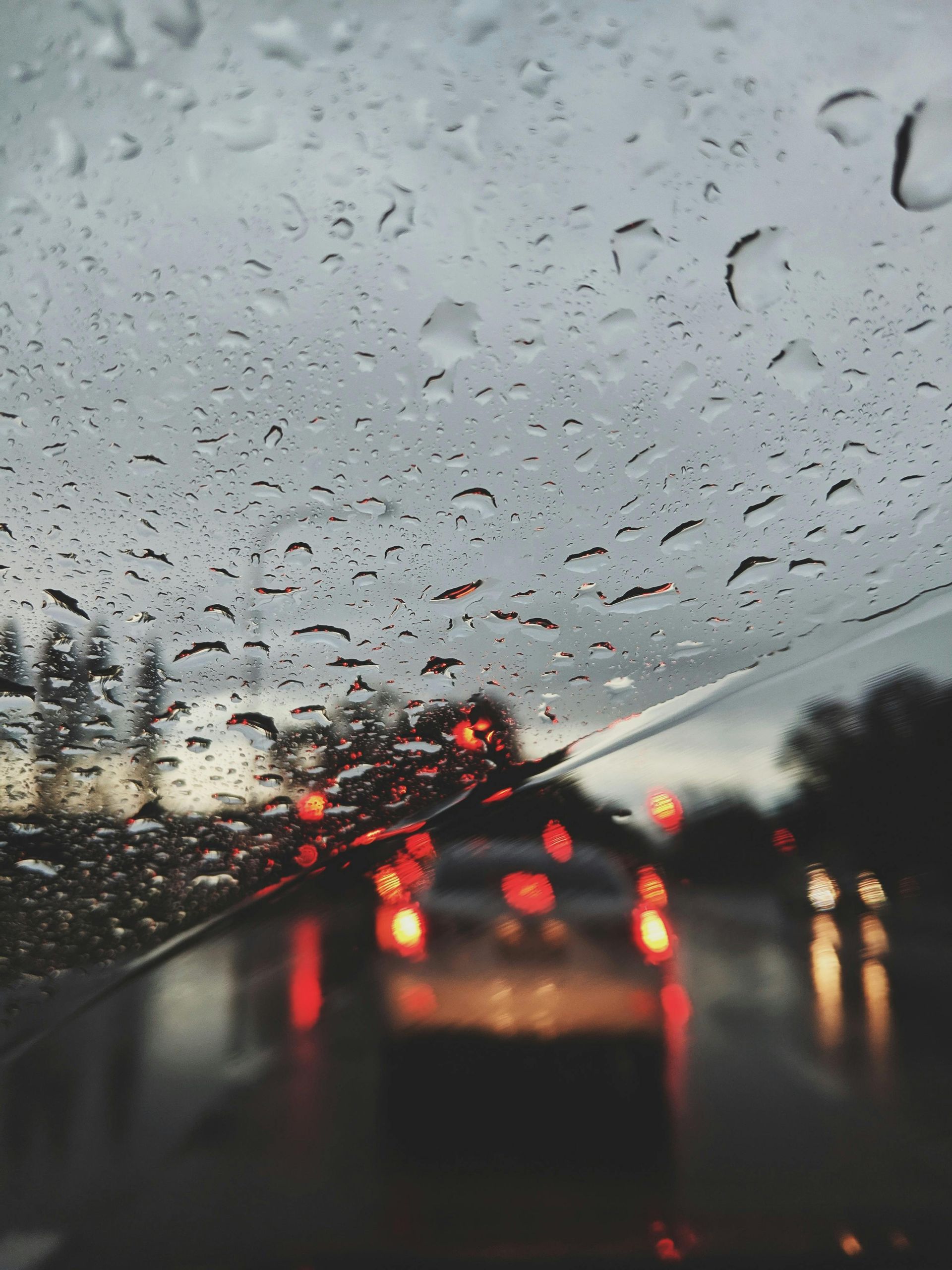 Raindrops on windshield, blurred view of traffic with red taillights in a dusky setting.