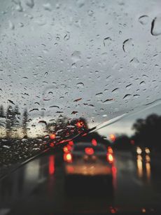 Raindrops on windshield, blurred view of traffic with red taillights in a dusky setting.