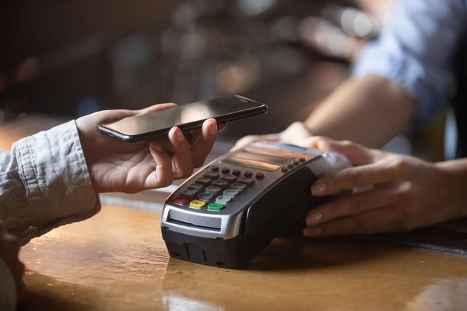 Una persona avvicina una carta a un terminale di pagamento mentre un'altra tiene uno smartphone sopra un bancone di legno.
