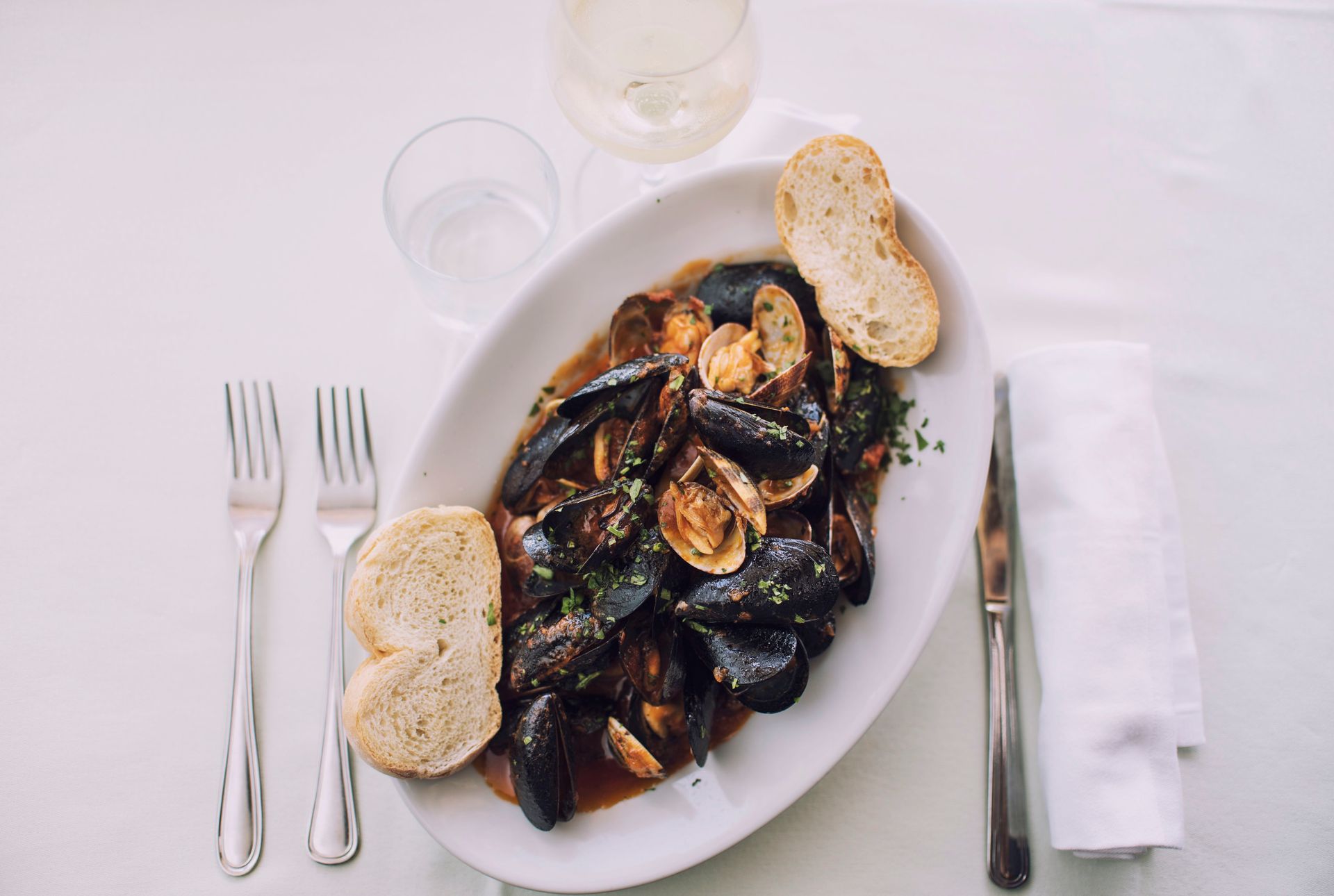 Cozze e vongole in salsa scura servite su un piatto ovale bianco con pane, posate e una bevanda.
