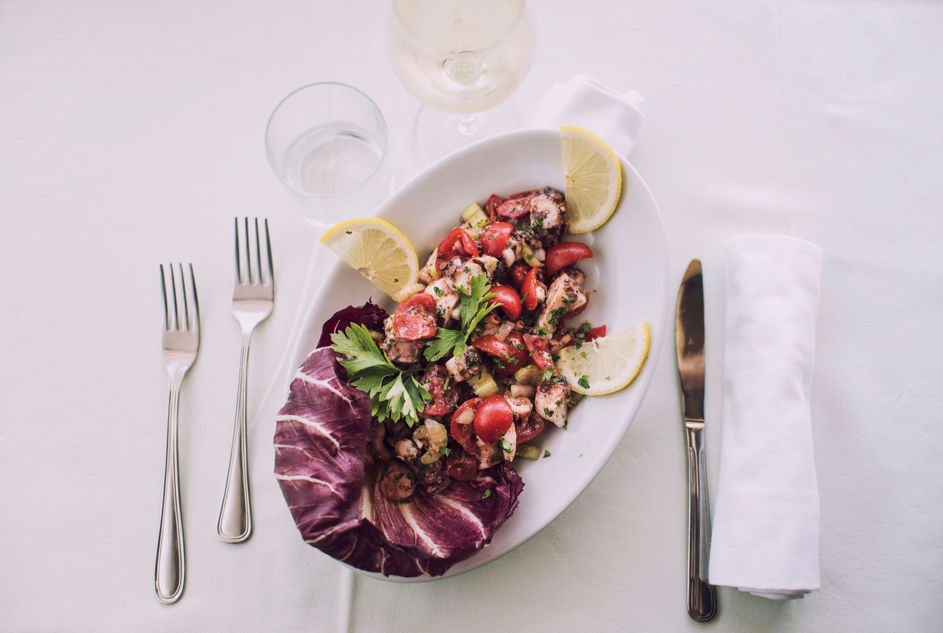 Insalata di polpo su un piatto ovale bianco con fette di limone, una forchetta, un coltello e una bevanda, il tutto su una tovaglia bianca.