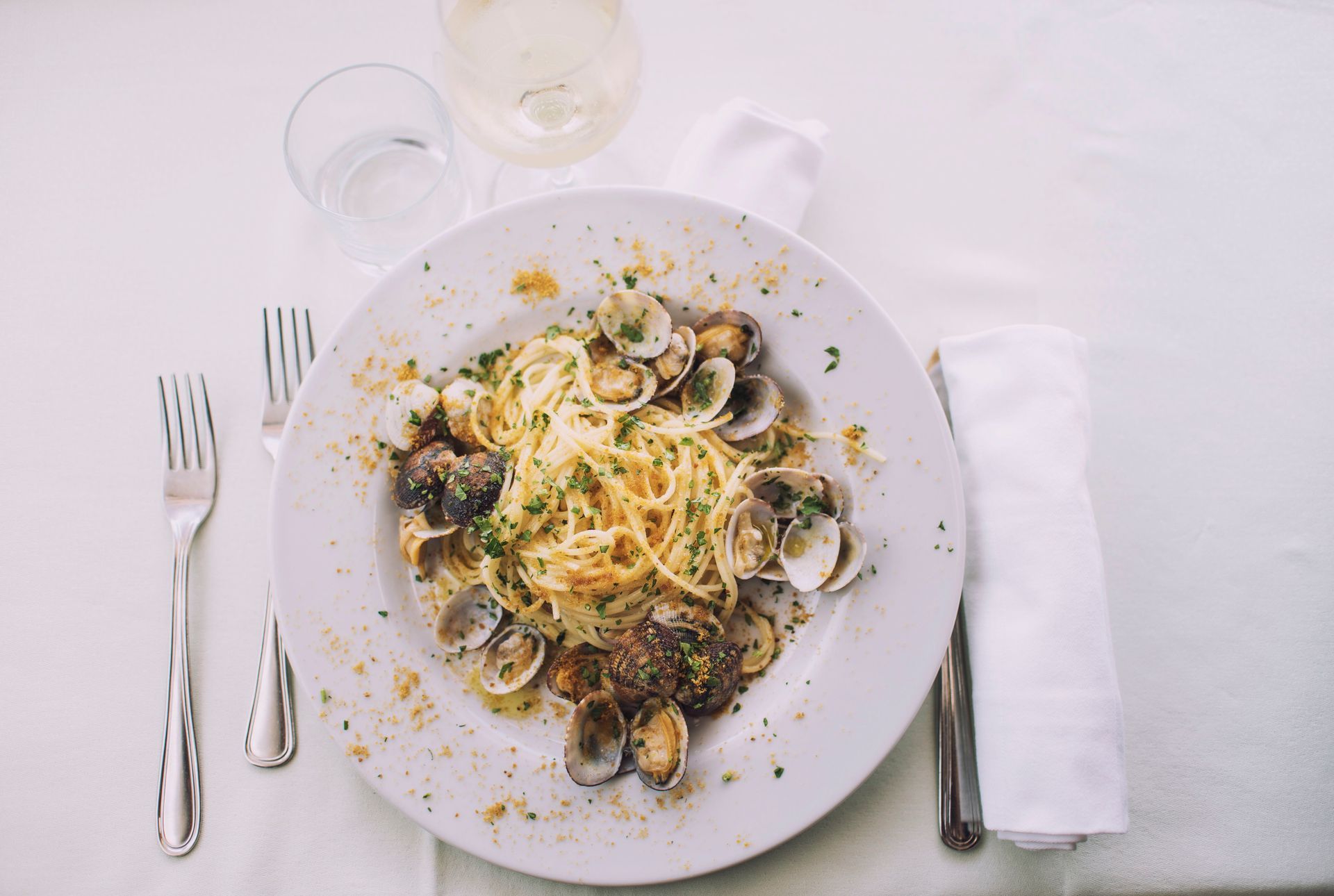 Spaghetti alle vongole su un piatto bianco, con posate e bevande su una tovaglia bianca.