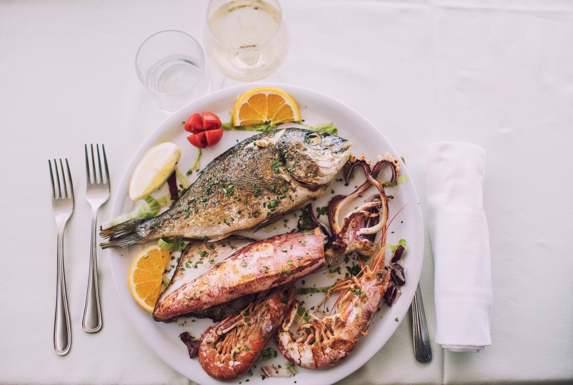 Piatto di frutti di mare: pesce alla griglia, gamberi, calamari, fette di limone e arancia su un piatto bianco con posate.