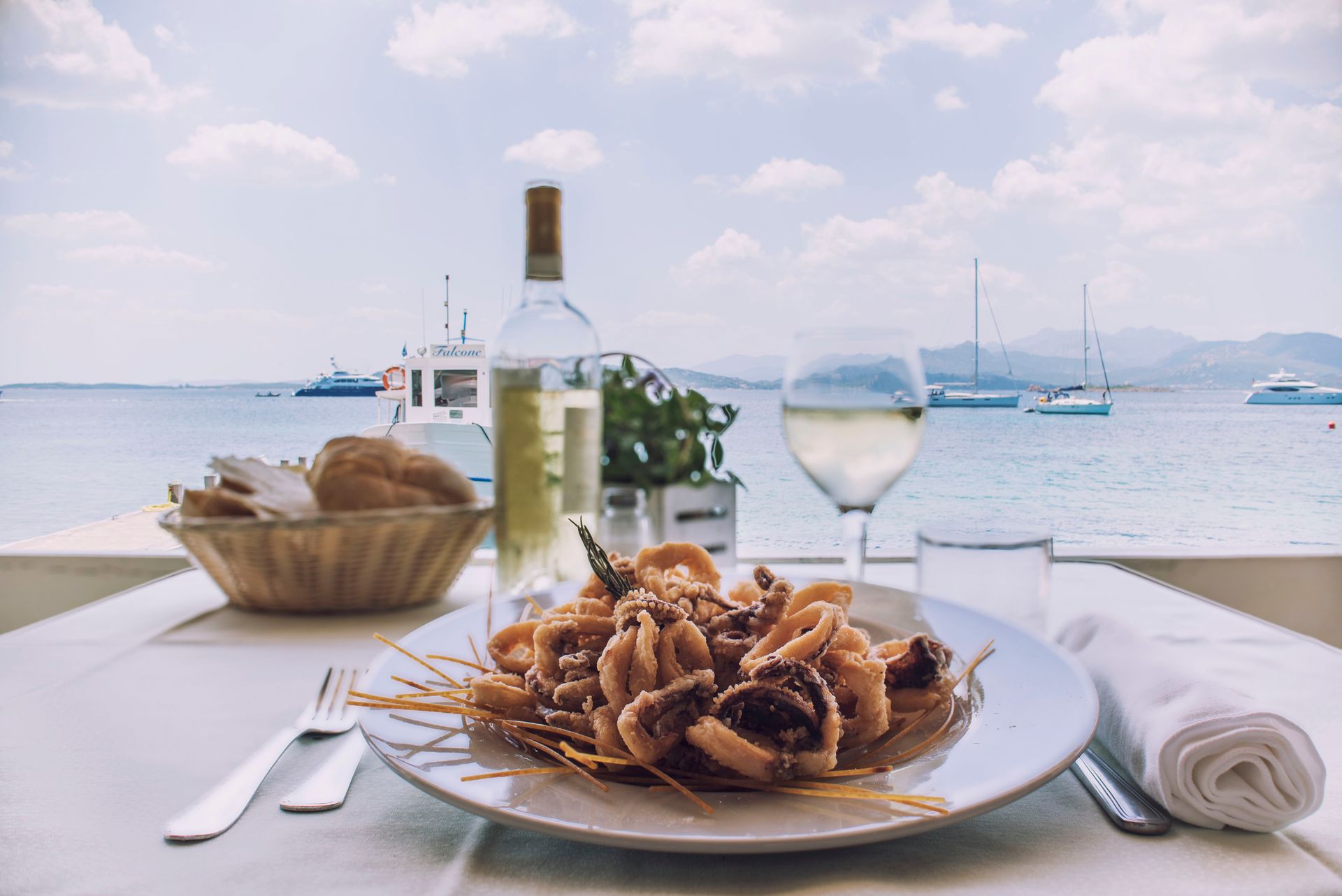 Tavolo con frutti di mare e vino, con vista su un mare azzurro e soleggiato, con barche.
