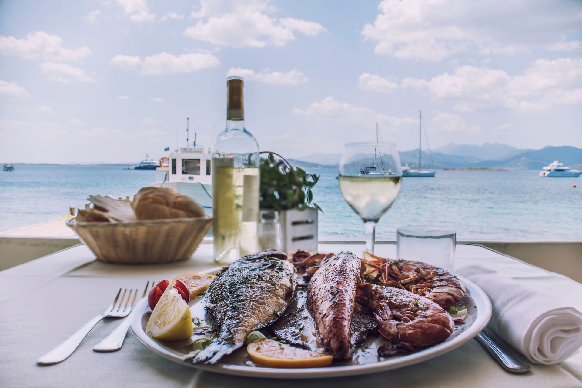 Piatto di frutti di mare, vino e pane su un tavolo con vista sul mare blu e sulle barche.