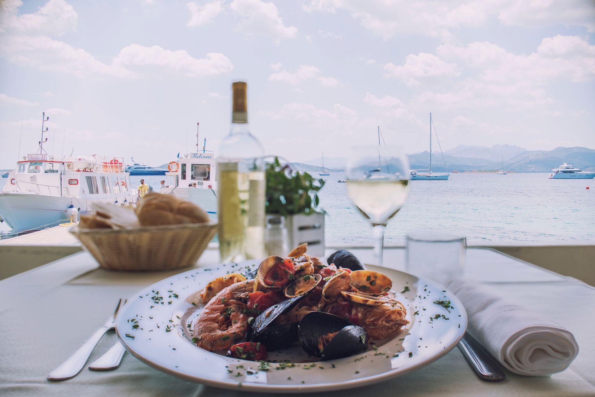 Piatto di frutti di mare con vino, pane e bicchiere d'acqua su un tavolo con vista sull'acqua e sulle barche sotto un cielo luminoso.