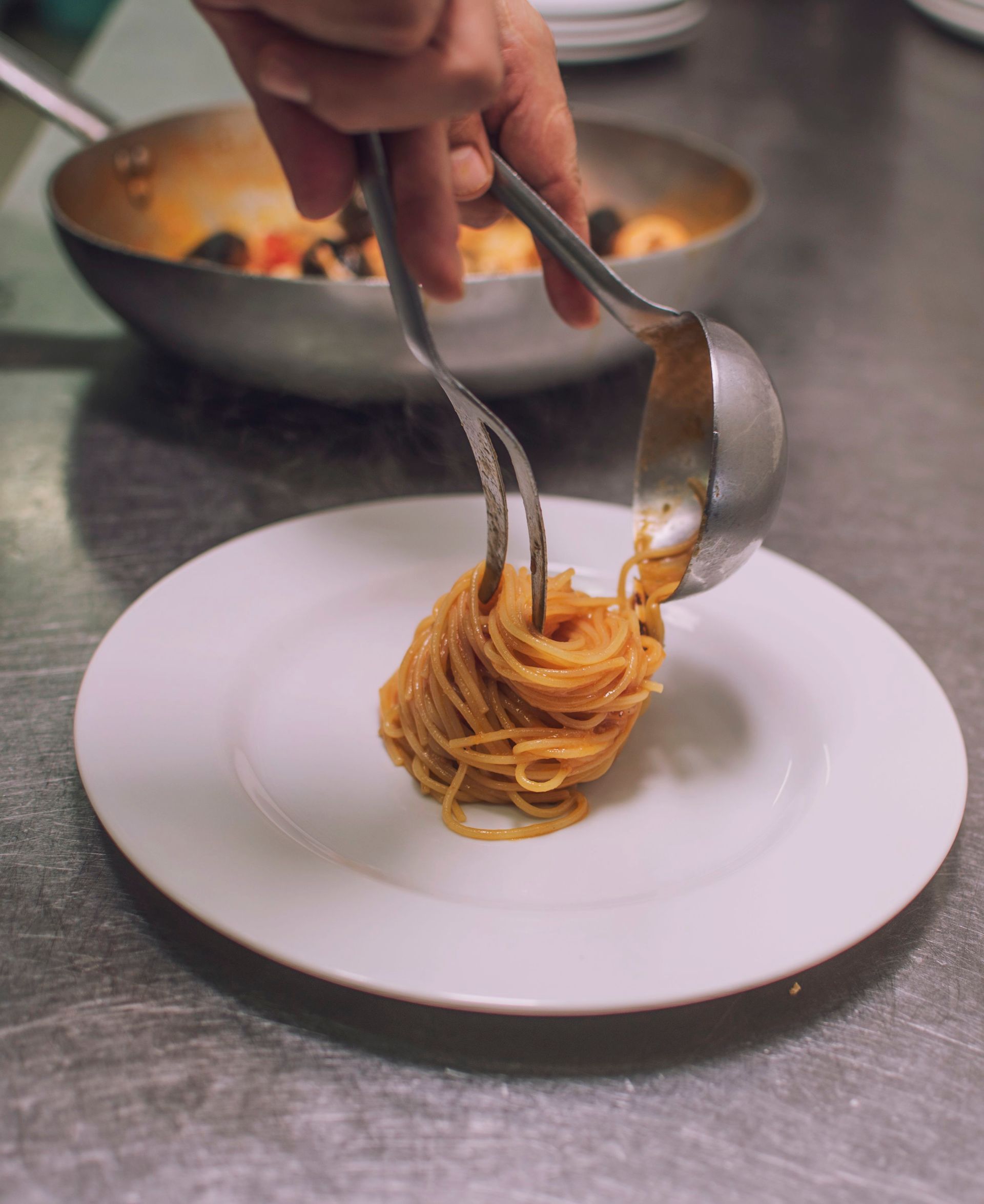Uno chef che impiatta gli spaghetti con pinze e mestolo, sullo sfondo salsa e padella.