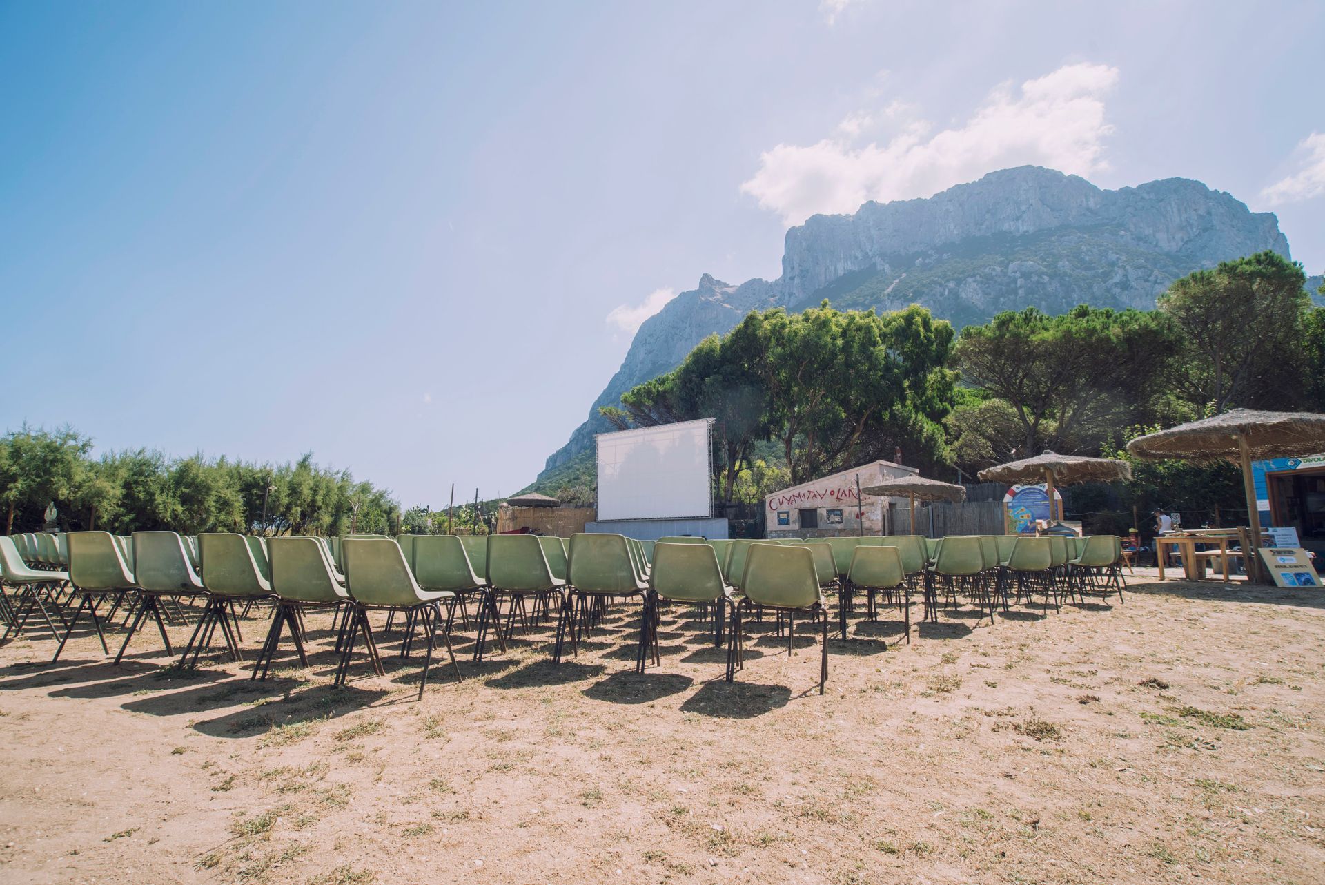 Cinema all'aperto sulla spiaggia; sedie verdi vuote di fronte a uno schermo bianco. Montagne sullo sfondo, giornata di sole.