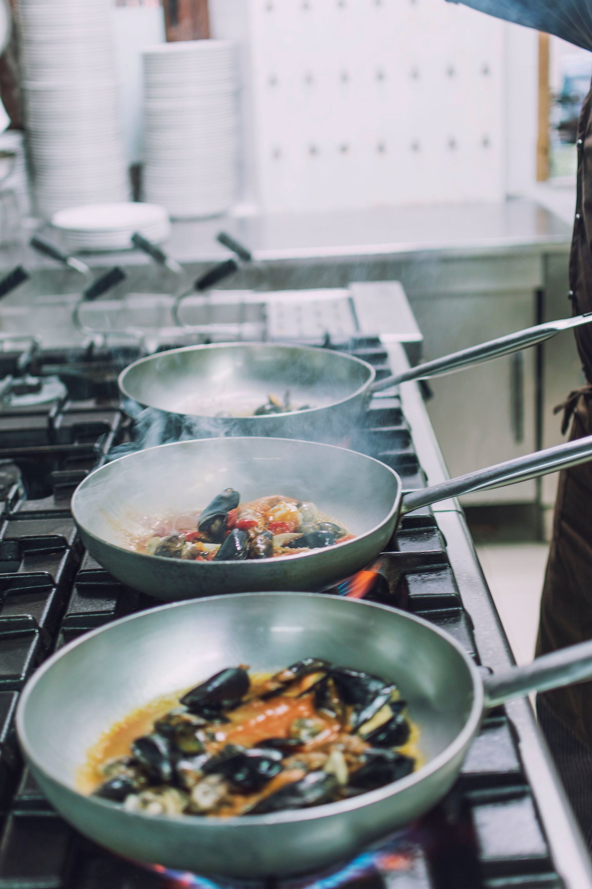 Tre padelle di pesce cuociono su un fornello a gas nella cucina di un ristorante. Il vapore sale.