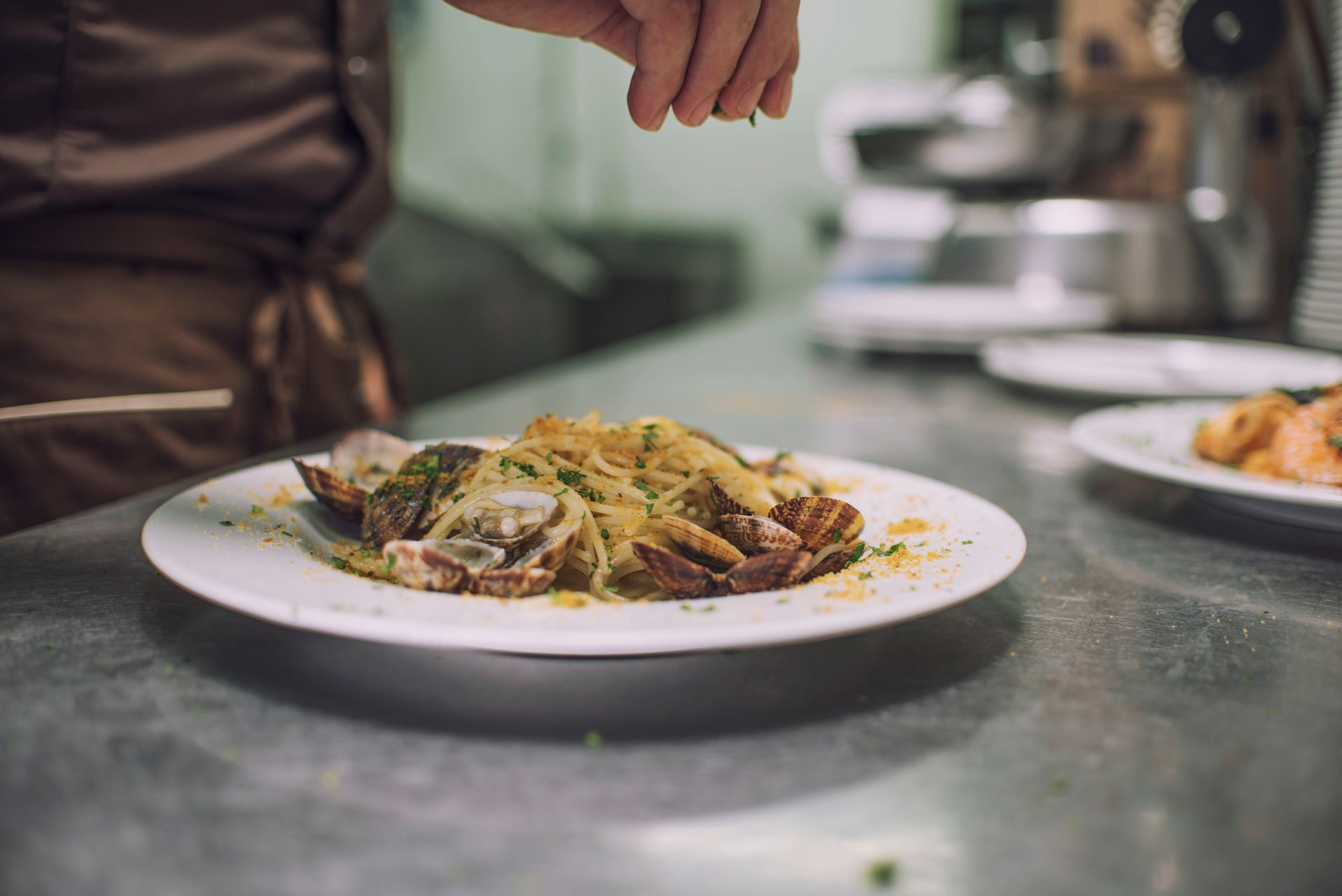 Lo chef guarnisce un piatto di pasta con erbe aromatiche su un piano di lavoro in acciaio inossidabile.