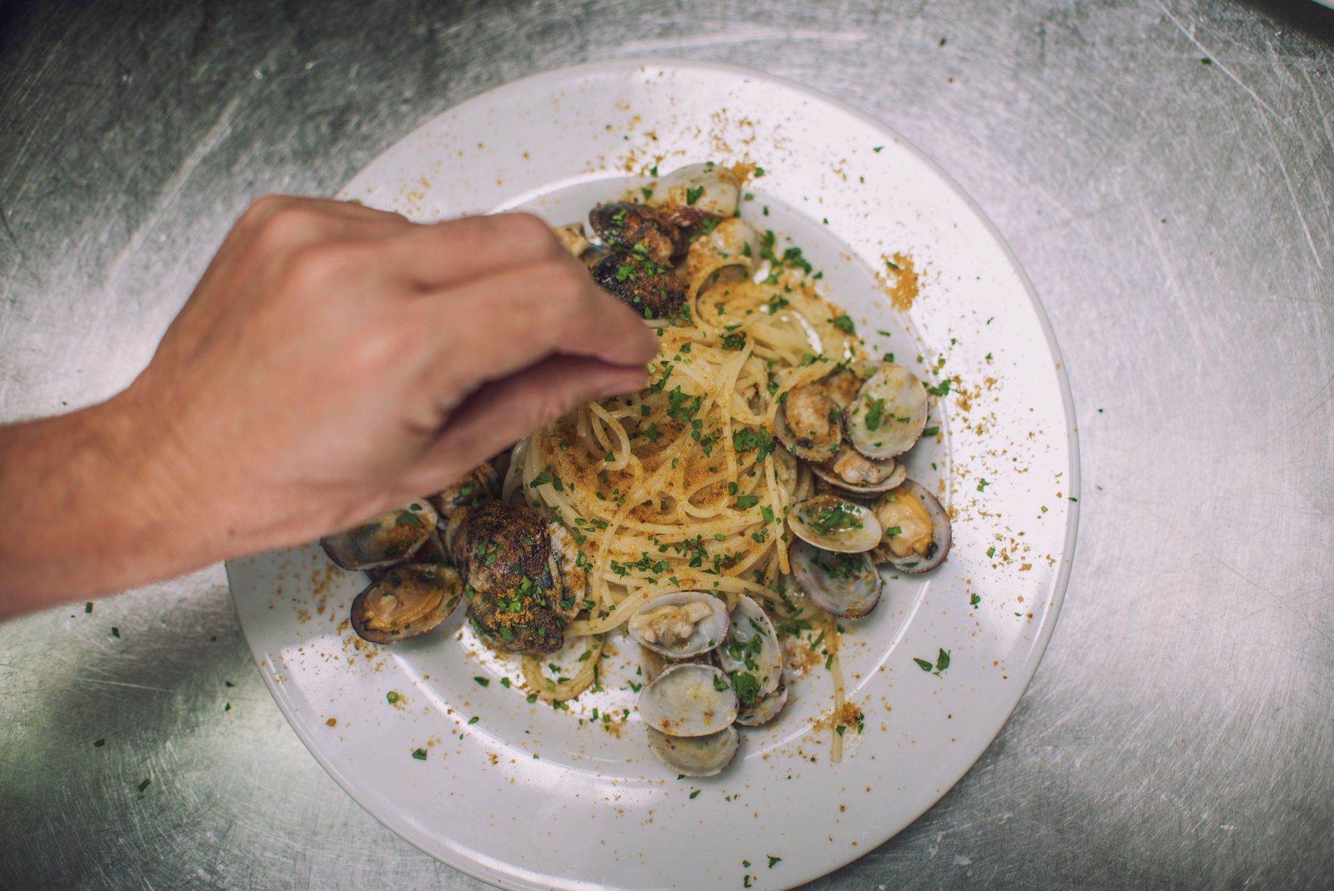 Una mano che cosparge di prezzemolo gli spaghetti alle vongole su un piatto bianco.