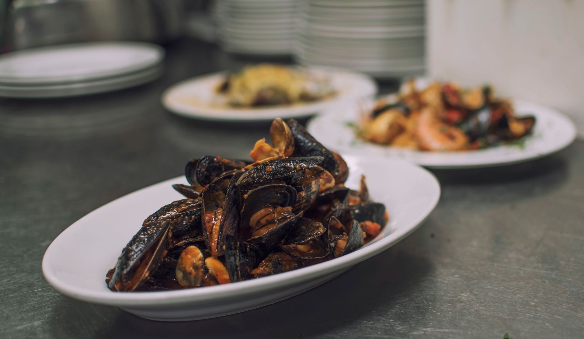 Primo piano di un piatto ovale bianco pieno di cozze cotte su un bancone in acciaio inossidabile; piatti sullo sfondo.