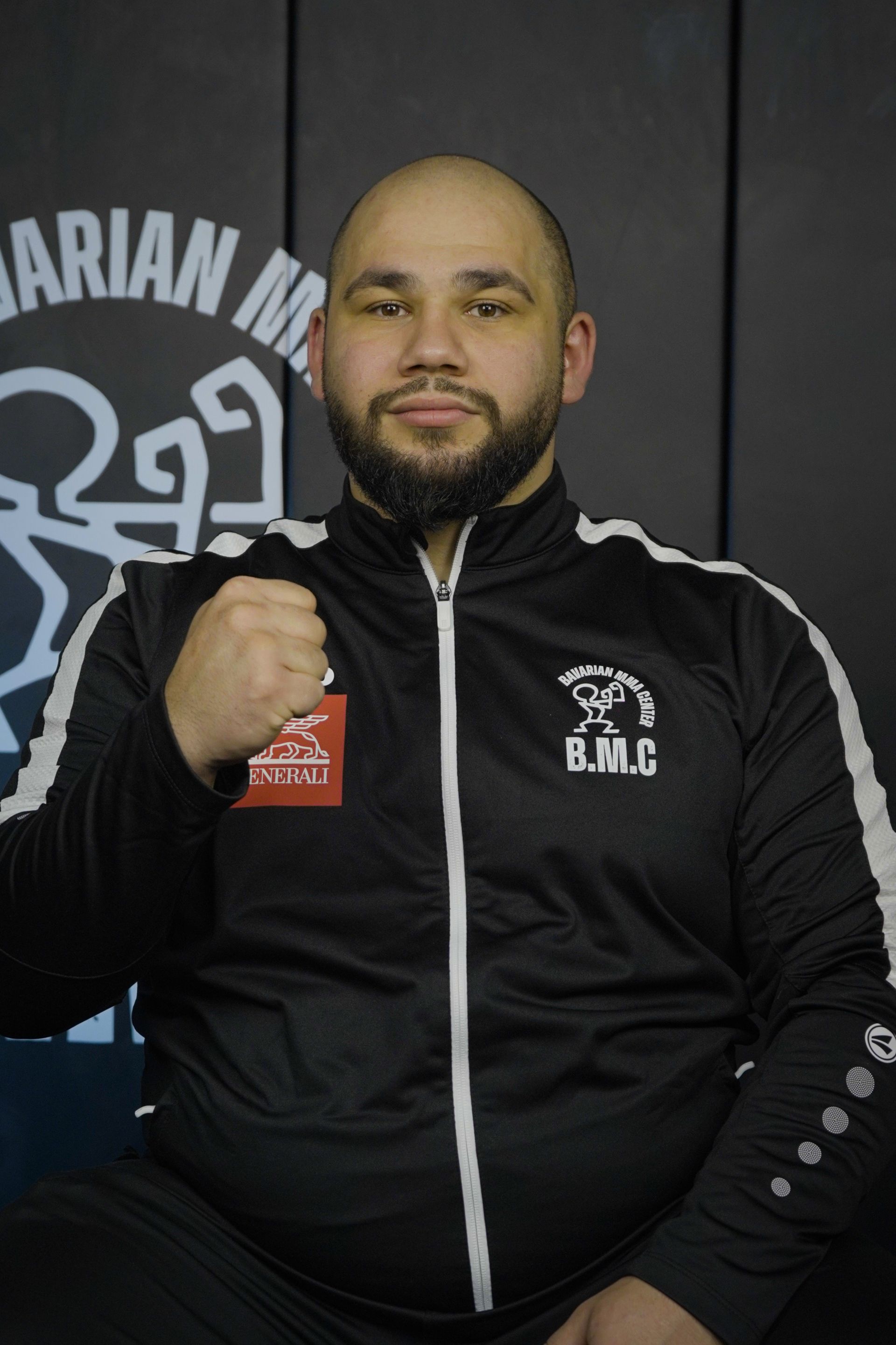 Ein Mann in einem schwarzen T-Shirt mit erhobenen Fäusten posiert vor einem Banner des Bavarian MMA Center.