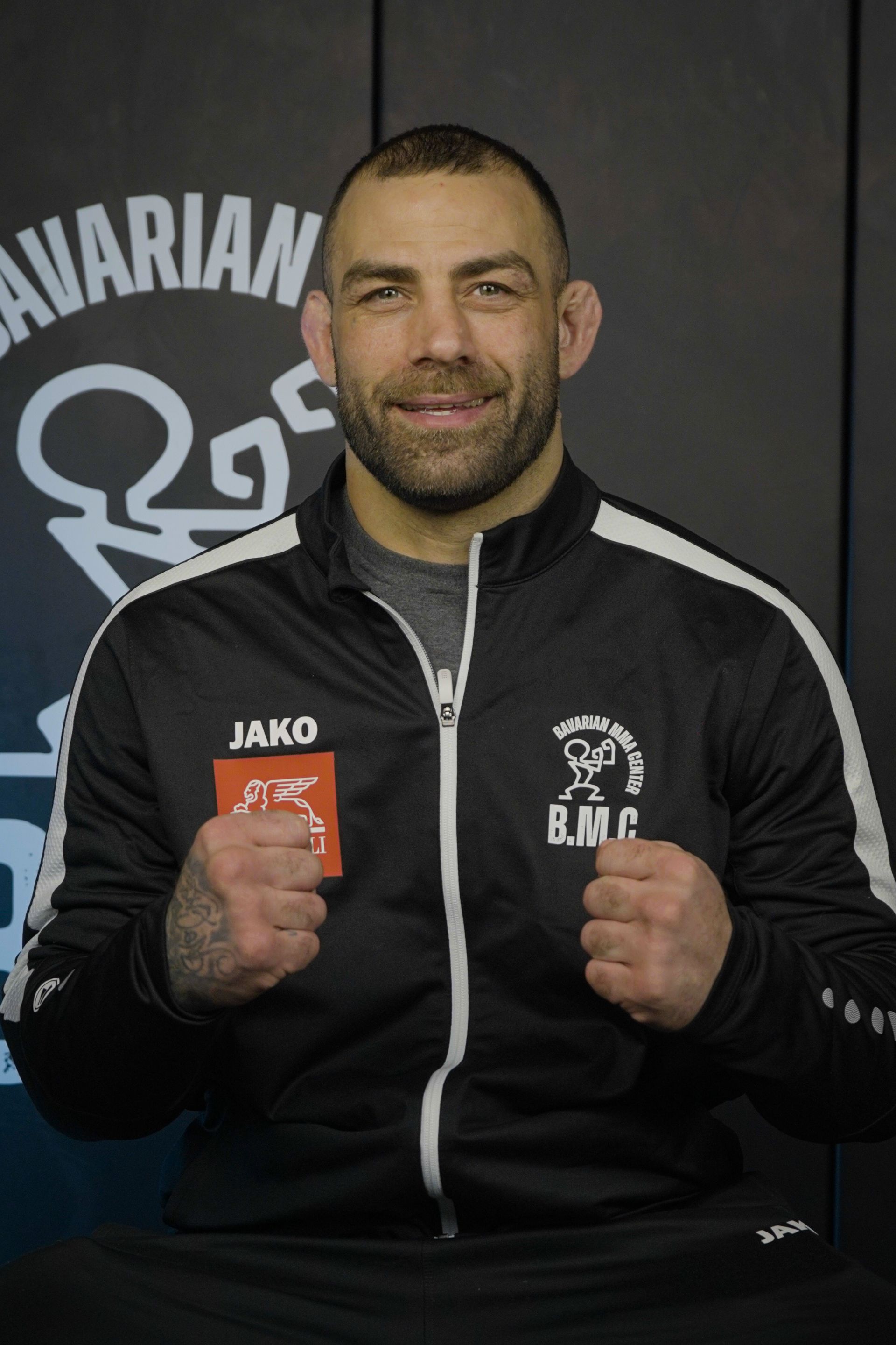 Ein Mann in einem grauen Langarmhemd gestikuliert mit den Händen und blickt nach unten. Er befindet sich in einer Turnhalle; im Hintergrund ist ein Spiegel zu sehen.