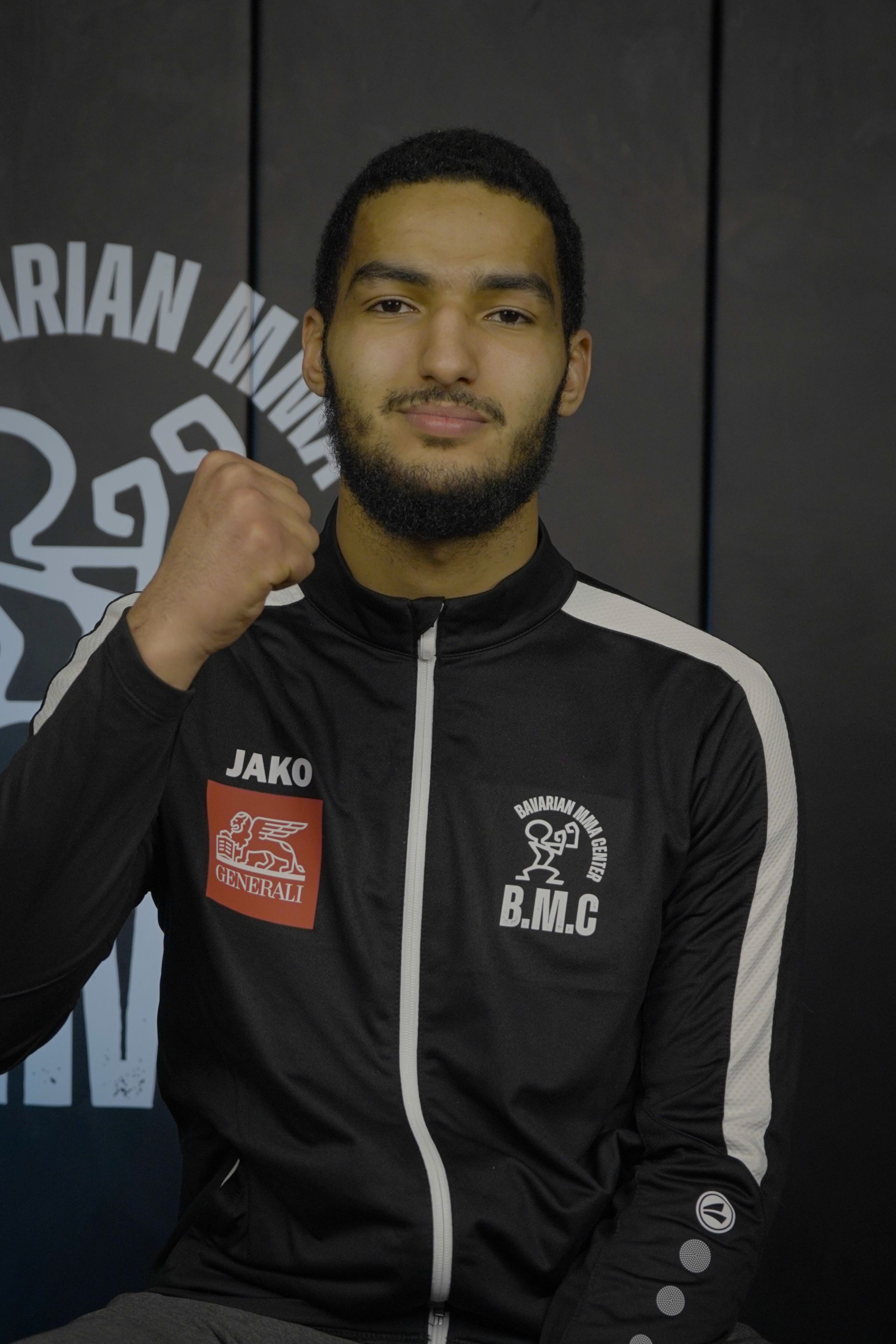 Ein Mann in einem schwarzen Kapuzenpulli steht vor einer Wand mit der Aufschrift „Bavarian MMA Center“.