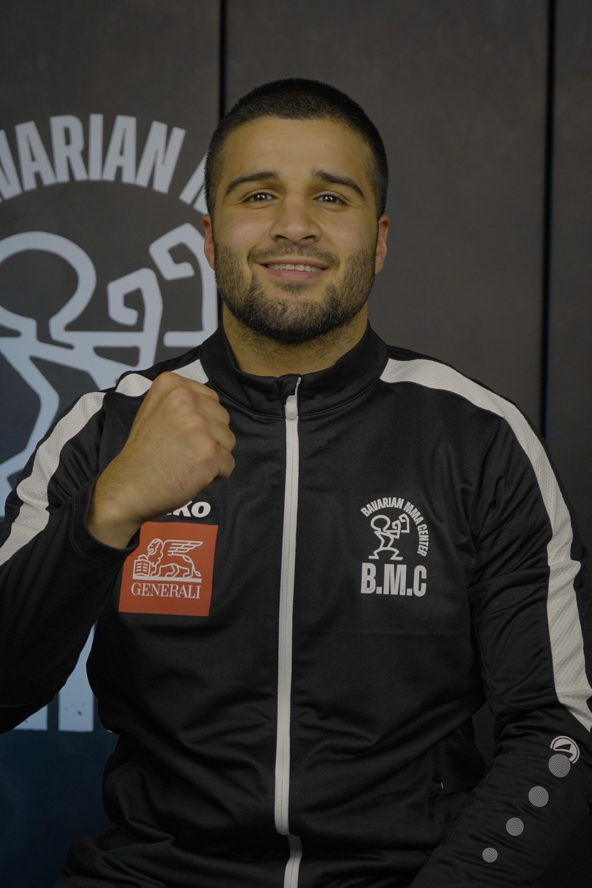 Ein Mann im blauen Hemd lächelt mit geballter Faust. Er steht vor dem Logo eines Fitnessstudios mit der Aufschrift „Bavarian MMA“.
