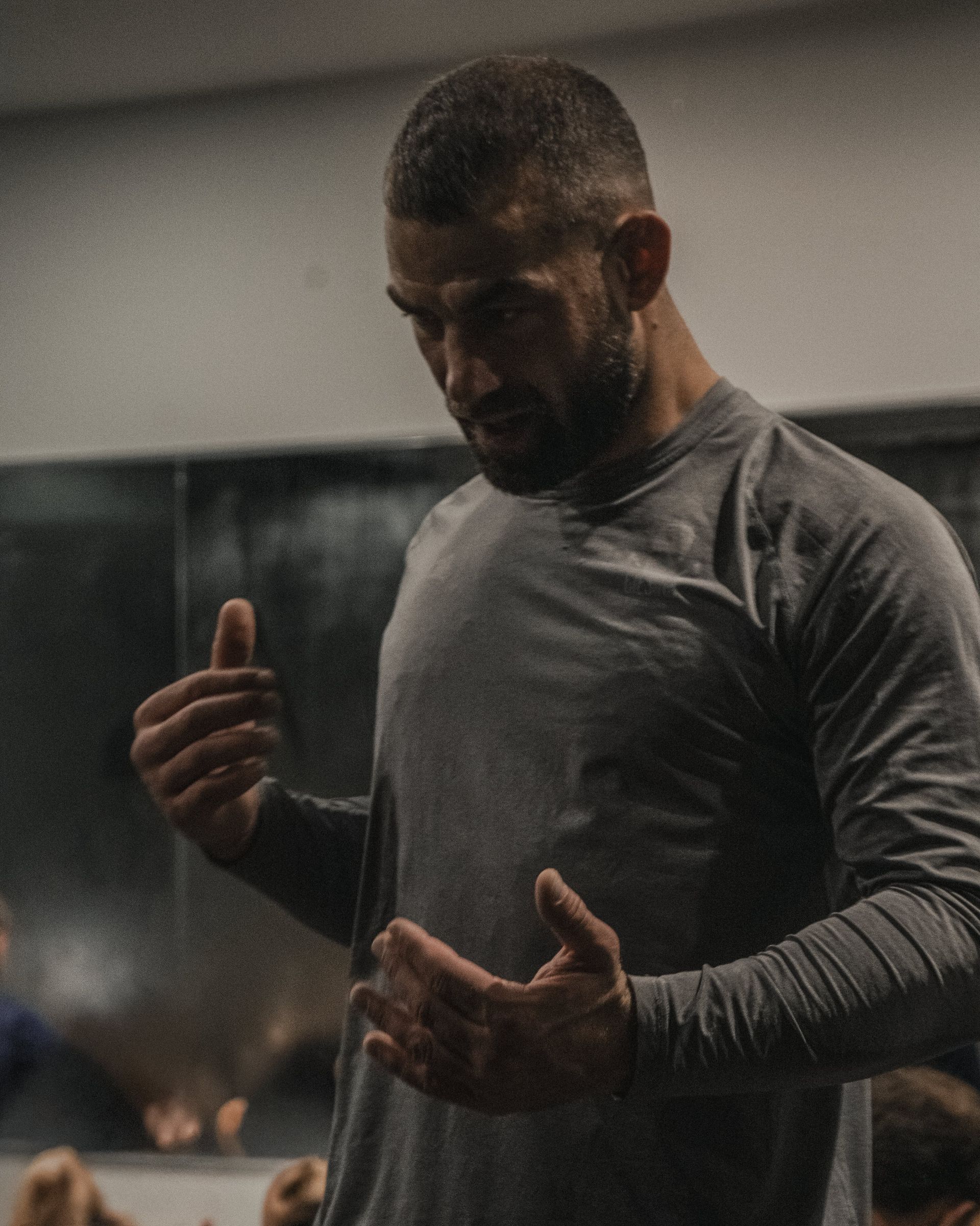 Ein Mann in einem grauen Langarmhemd gestikuliert mit den Händen und blickt nach unten. Er befindet sich in einer Turnhalle; im Hintergrund ist ein Spiegel zu sehen.