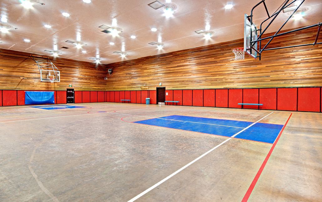 Indoor gym with a basketball court, red padded walls, and bright overhead lighting.
