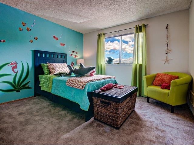 Bedroom in an apartment with a teal bed, ocean-wall mural, wicker trunk, and green curtains.
