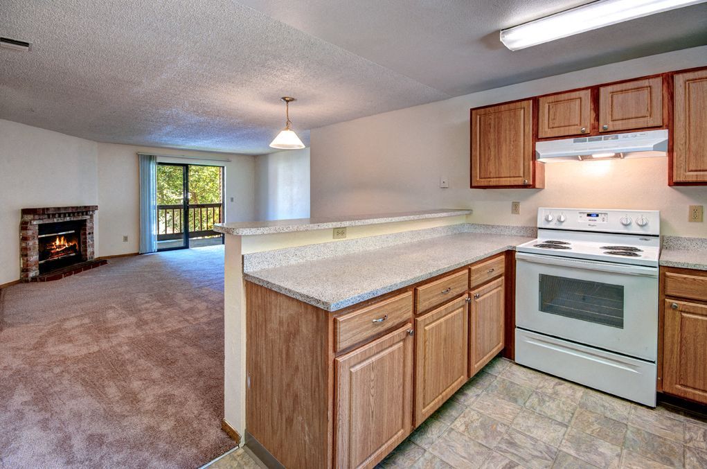Open-concept kitchen with wood cabinets, light speckled countertops, and a living area with a fireplace.