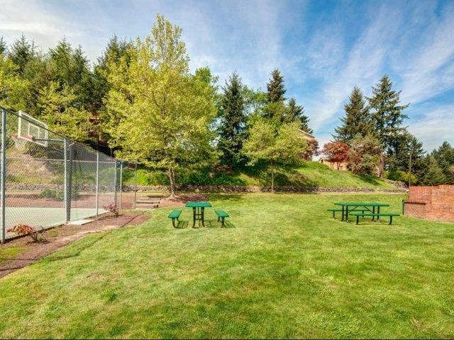 Open grassy outdoor community area with a tennis court fence, picnic tables, and trees under a blue sky.