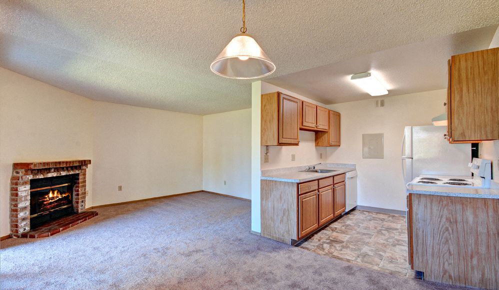 Living room with a brick fireplace on the left and an open kitchen with wood cabinets.