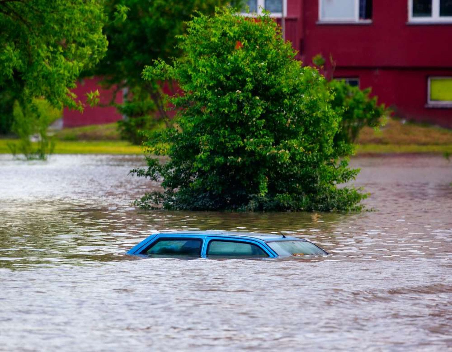 Flooded street; blue car submerged, green tree partially visible, red building in background.