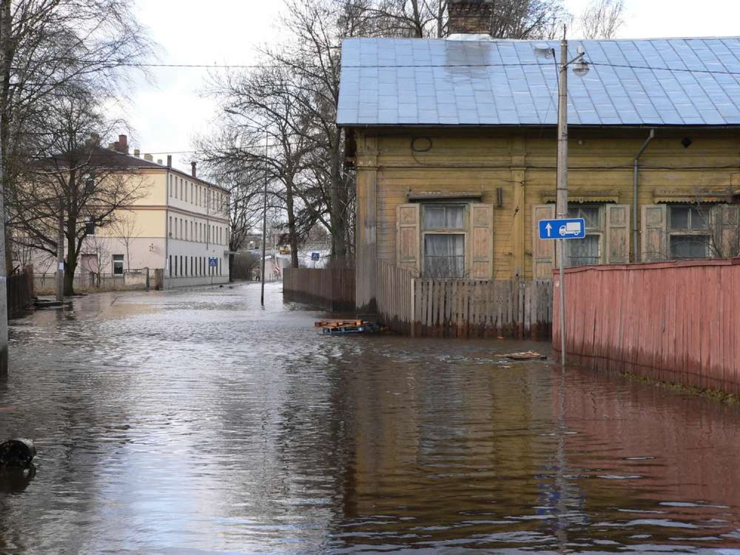 Flooded street with water covering the road, in front of buildings, under a cloudy sky.