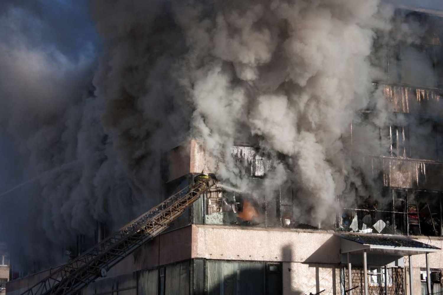Building engulfed in smoke and flames, with a fire truck ladder extended towards the building.