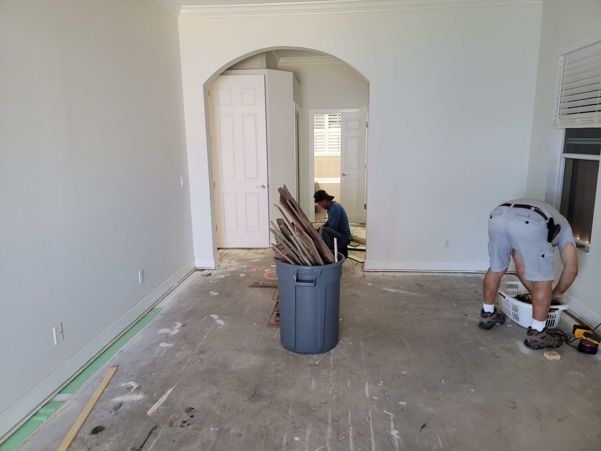Room under renovation, trash can of wood, people working. Light walls, arched doorway, open doorway.