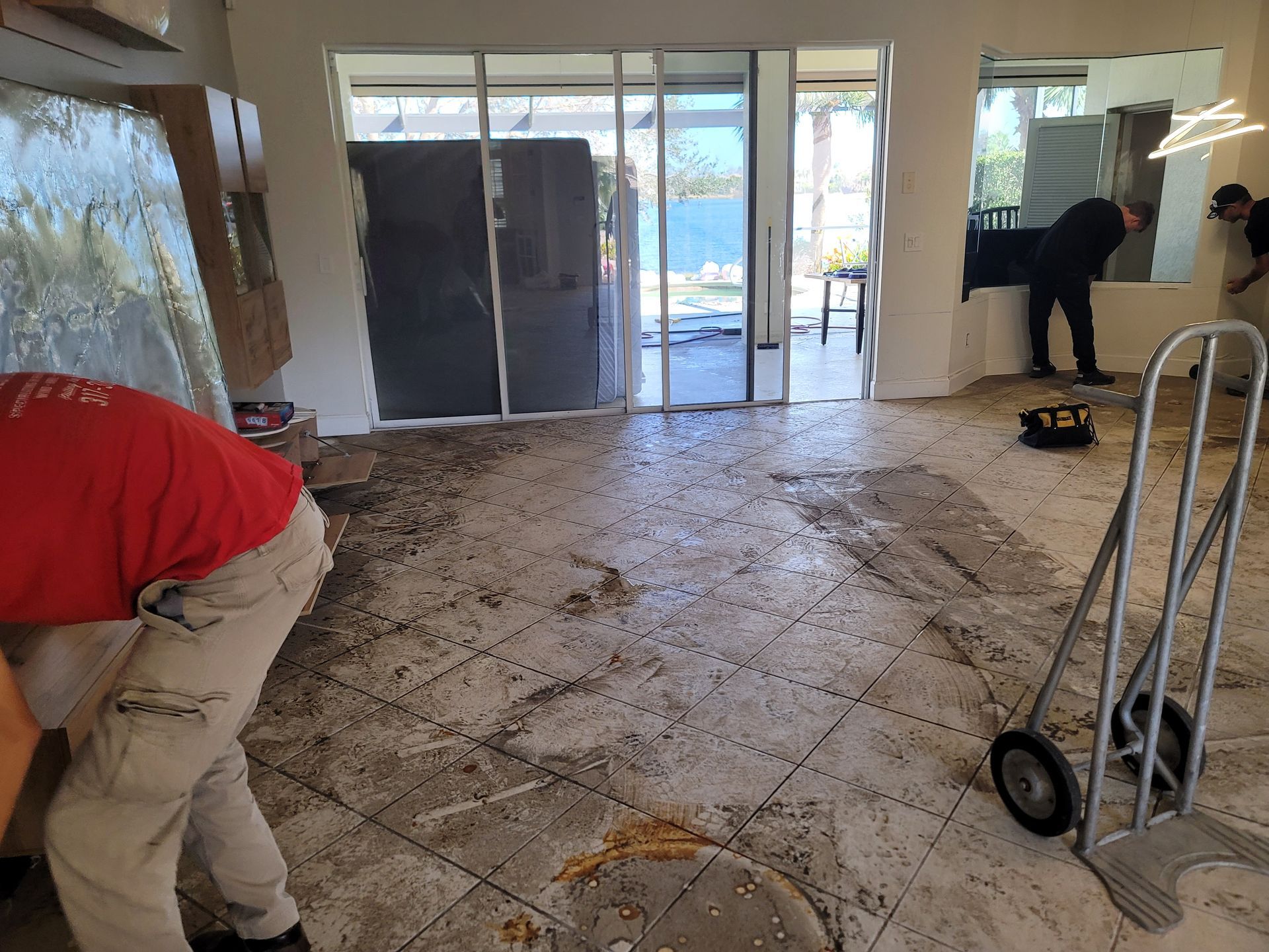 Workers removing tile flooring in a home, debris scattered. One man in red shirt bends down, hand truck present.
