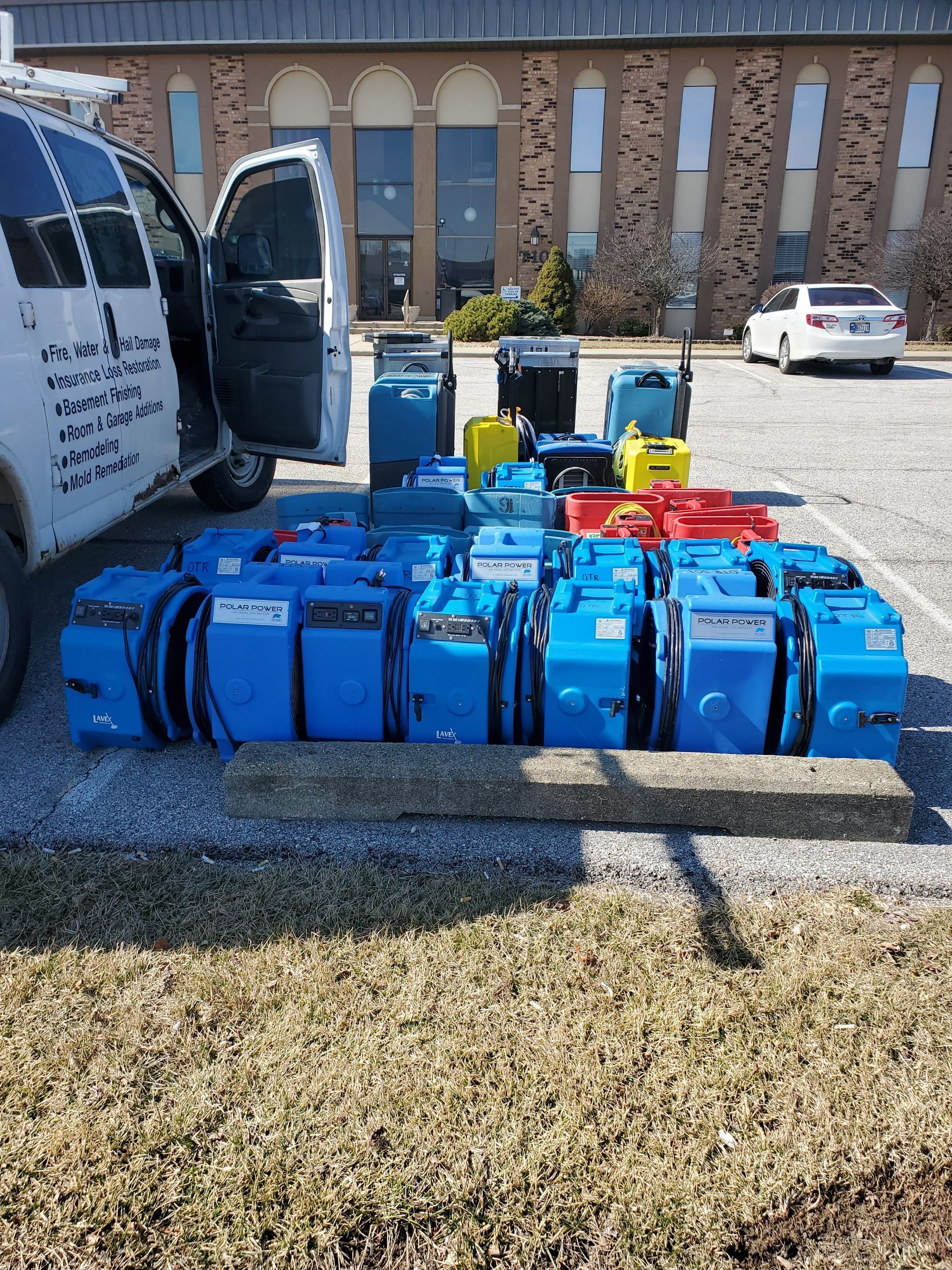 A van with open door parked next to many blue and colored electric floor cleaners on a concrete pad.