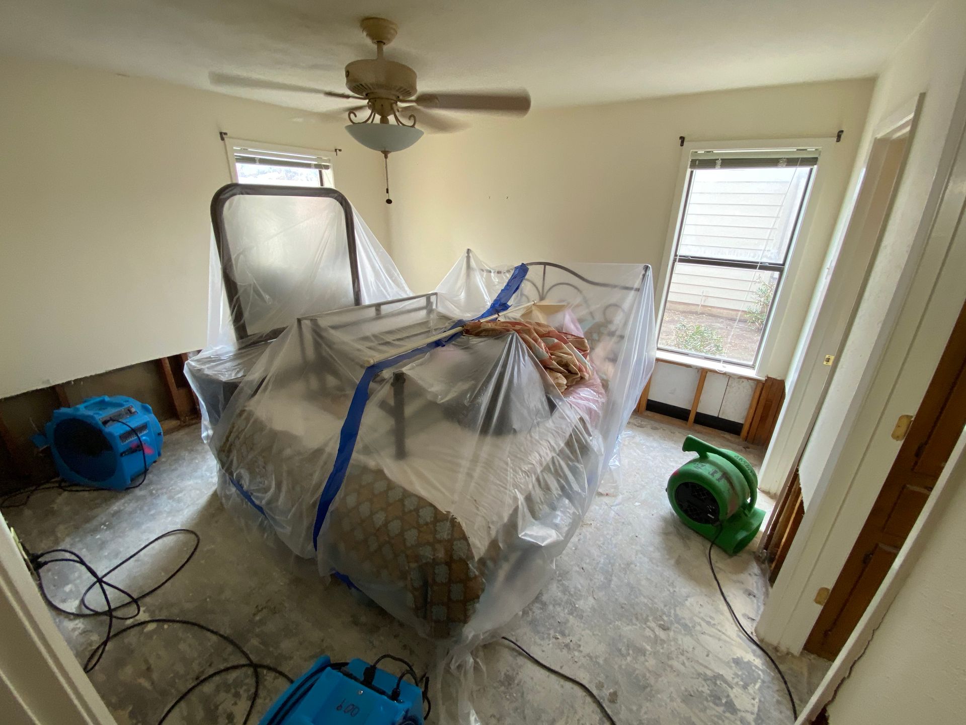 Bedroom with bed covered in plastic, two dehumidifiers, and exposed wall.