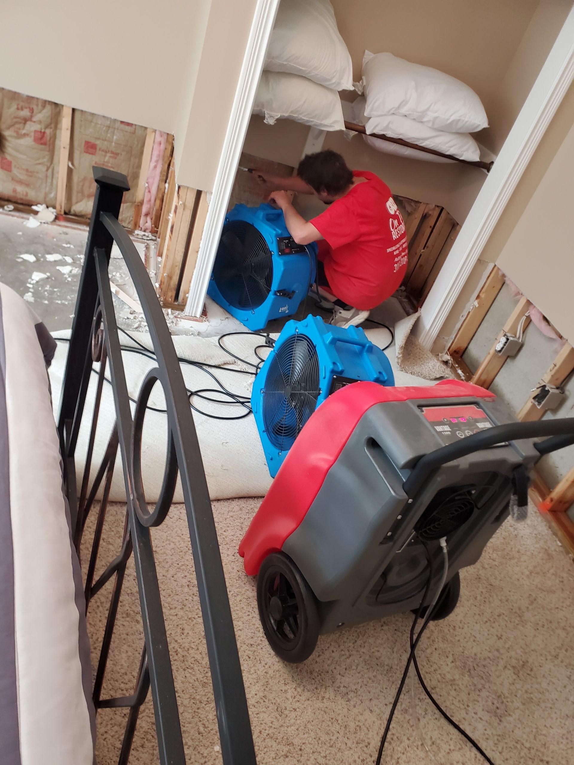 A person in red shirt sets up drying equipment in a flooded room, exposed wall, with two blue fans and a red dehumidifier.
