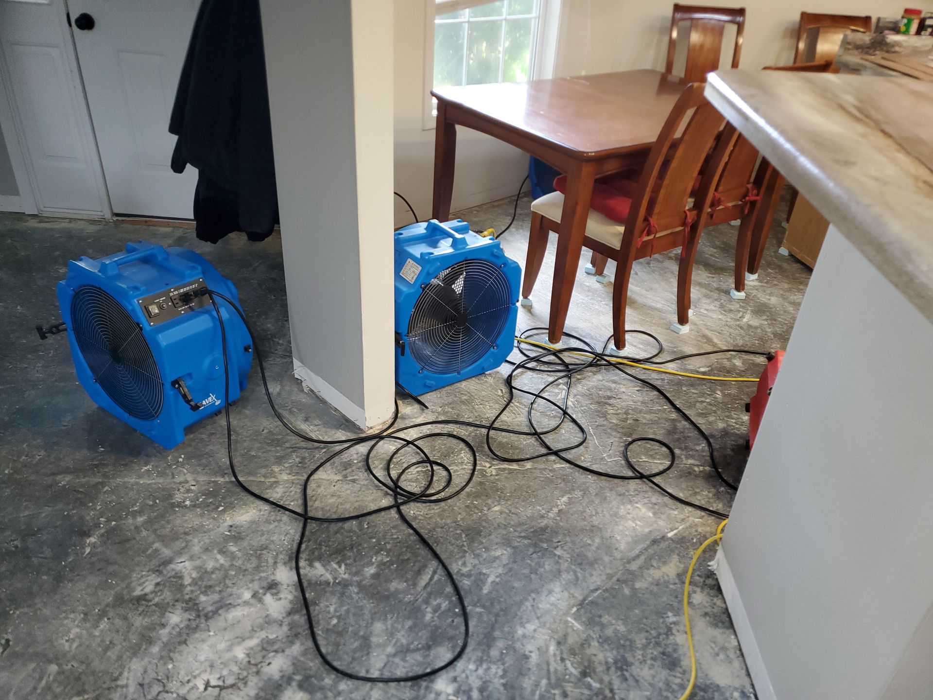 Two blue fans with black cords on a concrete floor drying a room, with a table and chairs in the background.