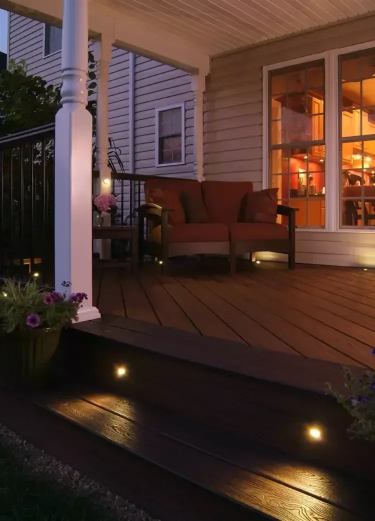 A dimly lit wooden porch at night, featuring a small loveseat, potted plants, and warm accent lighting on the steps.