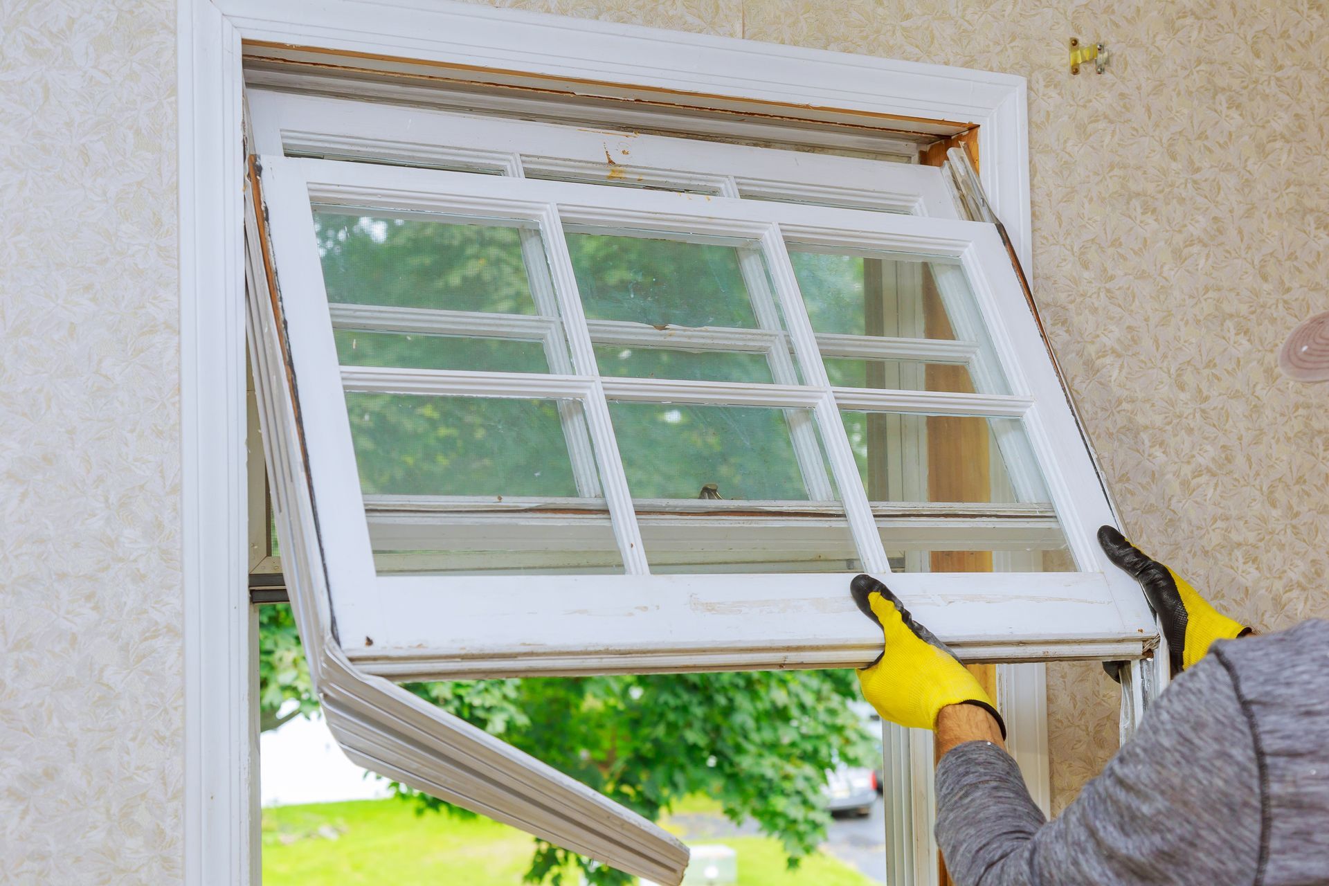 Person installing a white-framed window in a wall opening, holding it with yellow gloves. Person installing a white-framed window in a wall opening, holding it with yellow gloves.