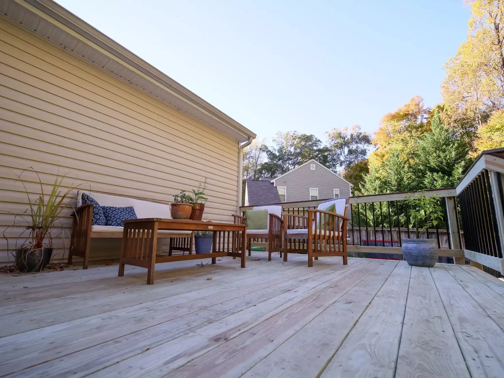 A wooden backyard deck with a couch, table, and chairs against a beige house wall under a clear blue sky.