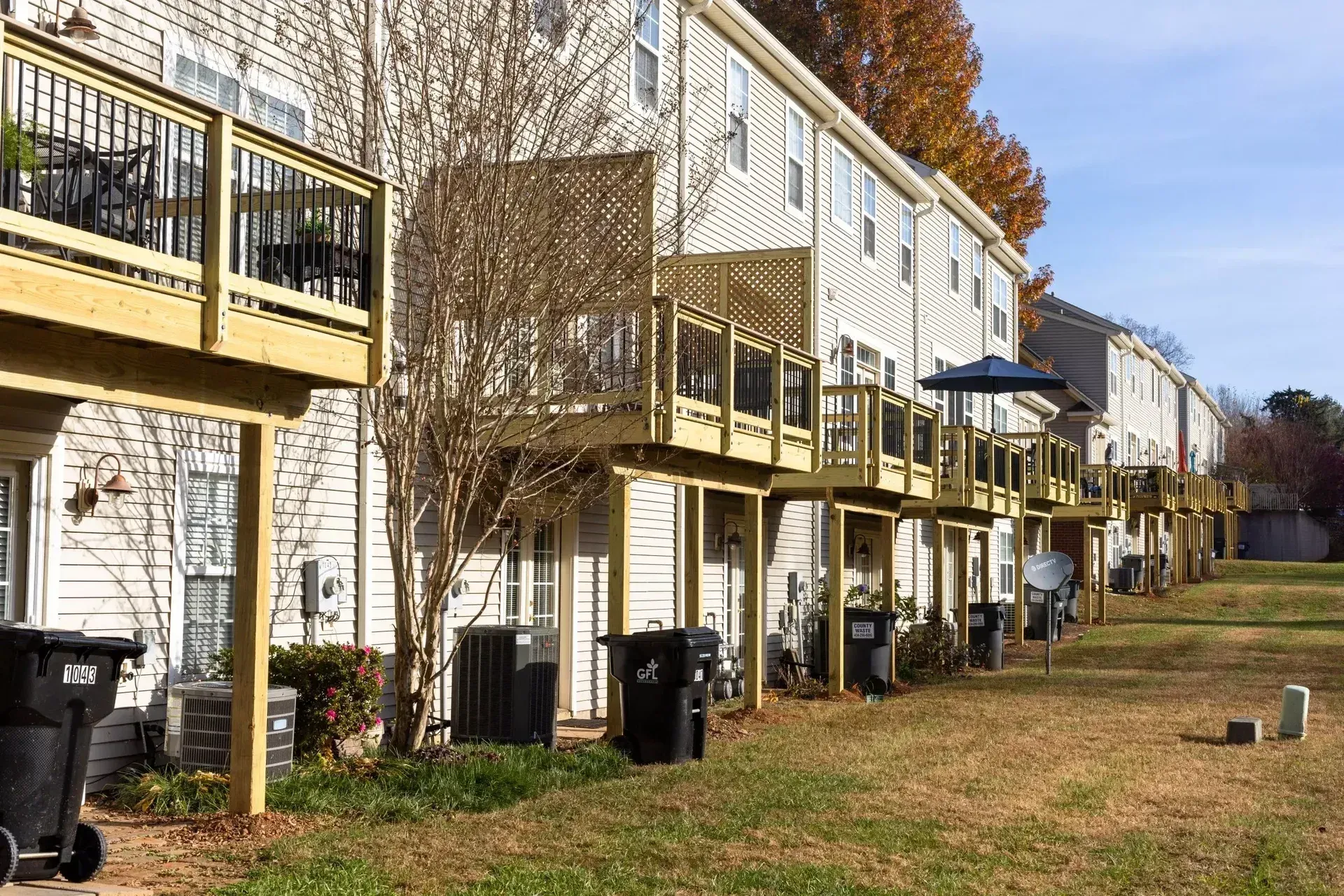 A row of multi-story townhomes with wooden elevated balconies, exterior staircases, and trash bins, seen from the side.