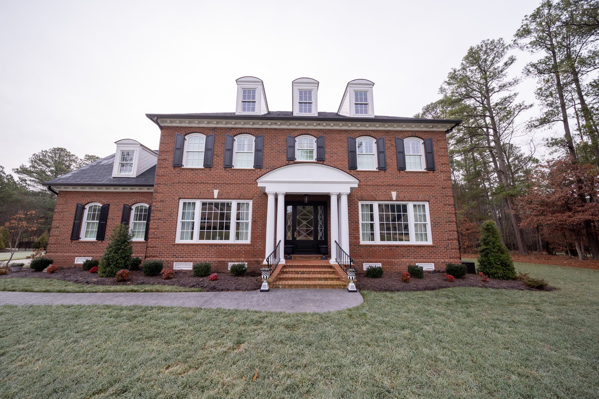 A two-story red brick colonial house with a white front portico, black shutters, and three dormer windows on the roof.