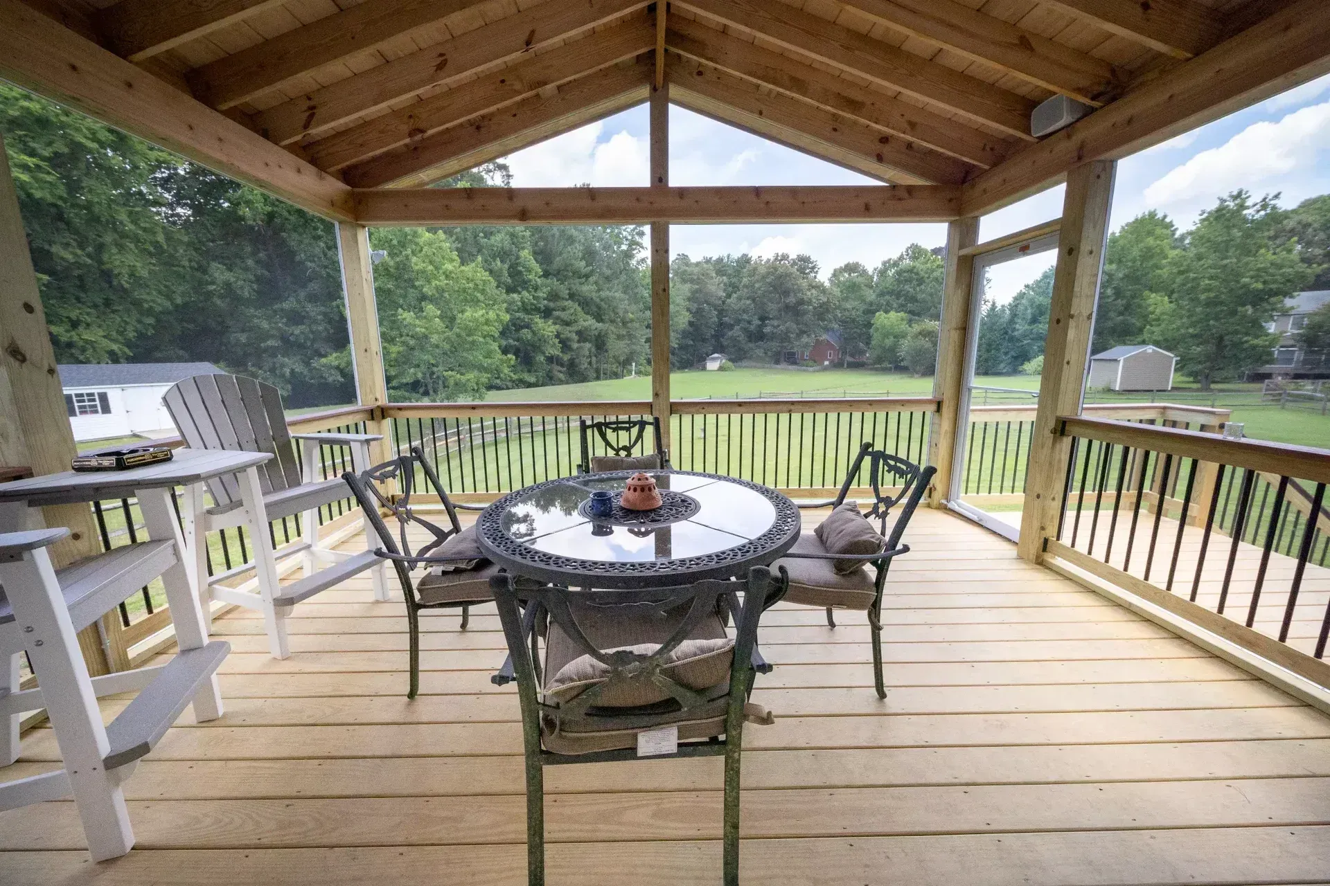 A covered wooden deck features a round glass-top table with metal chairs, overlooking a grassy backyard and trees.