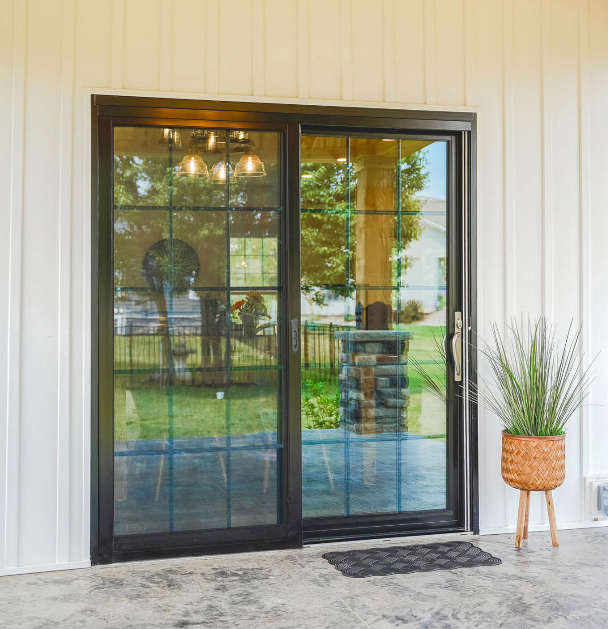 A wooden deck with a railing leads to sliding glass doors on the side of a gray-sided house with a large overhang.