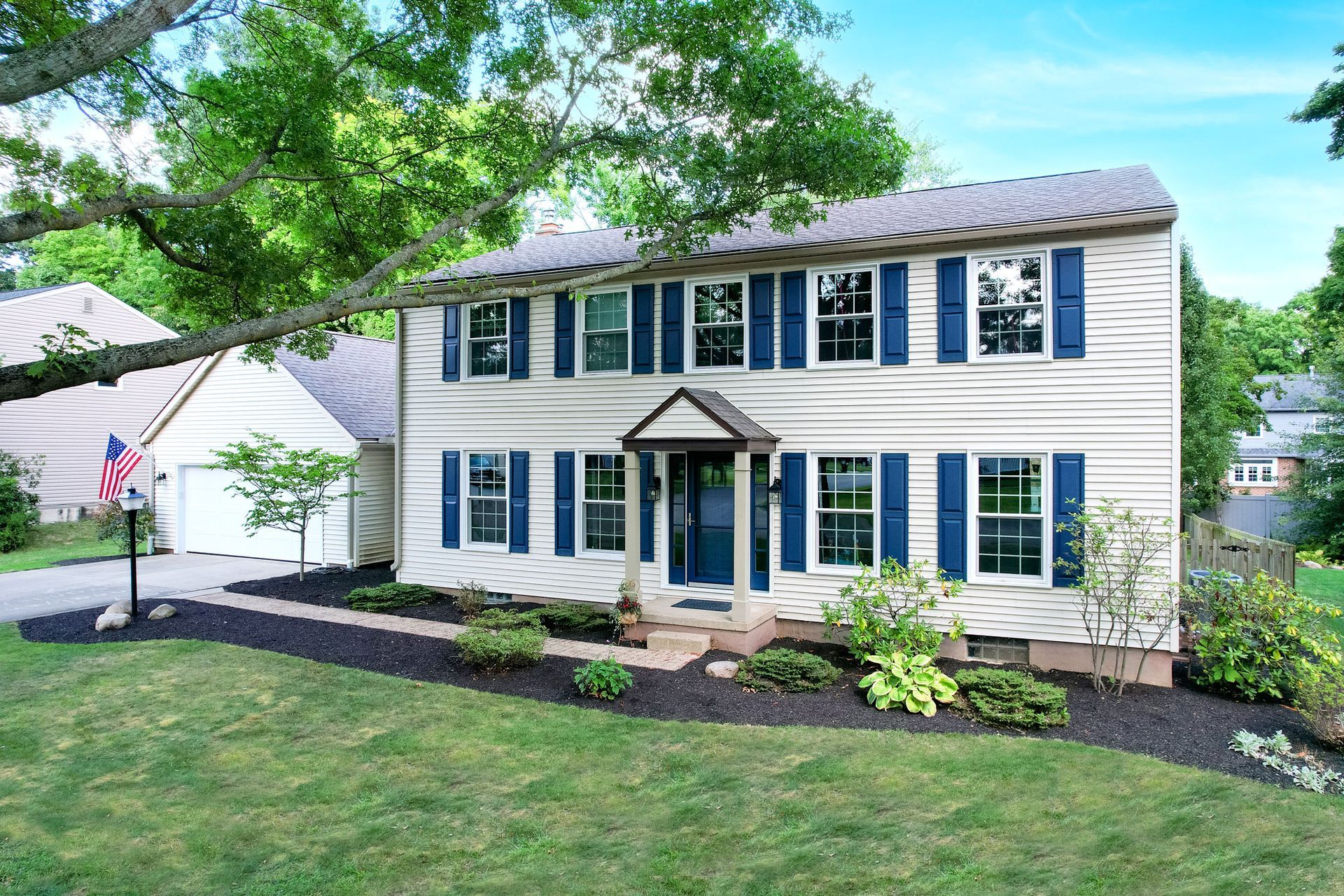 A two-story colonial house with beige siding, blue shutters, a dark roof, and a small front porch, set in a green yard.
